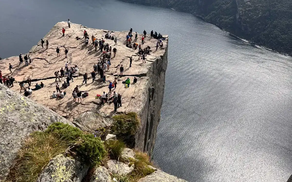 Dozens of people on a flat rock plateau high above the shining water of a fjord