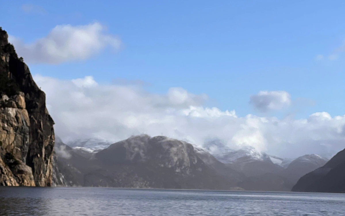 Fjord water with a tall brown cliff on the left and snow-dusted peaks in white cloud ahead