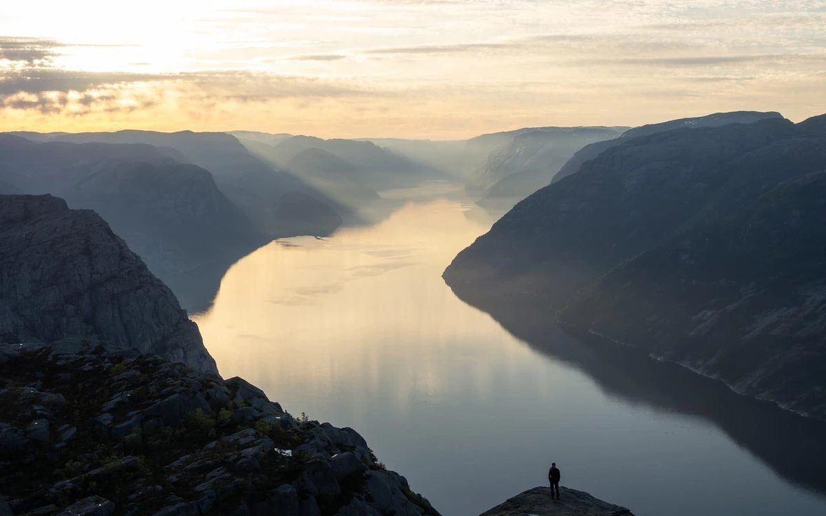 A hiker on a rocky ledge above the long Lysefjord with the low sun behind cloud