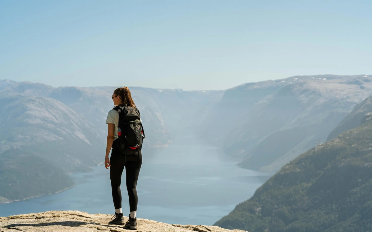 A hiker with a small backpack on bare rock looking down a long hazy fjord