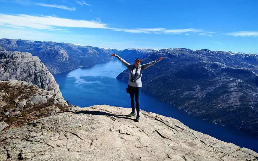 A hiker with arms spread on bare rock above a long blue fjord
