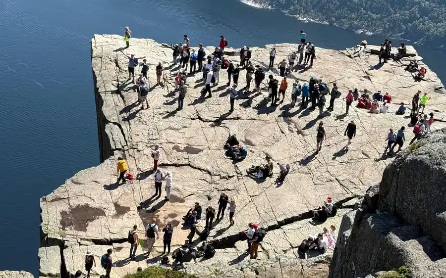 Many people standing and sitting on a wide flat rock above a fjord