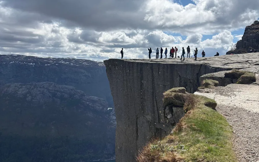 A line of hikers on the edge of a flat, sheer-sided cliff above a fjord