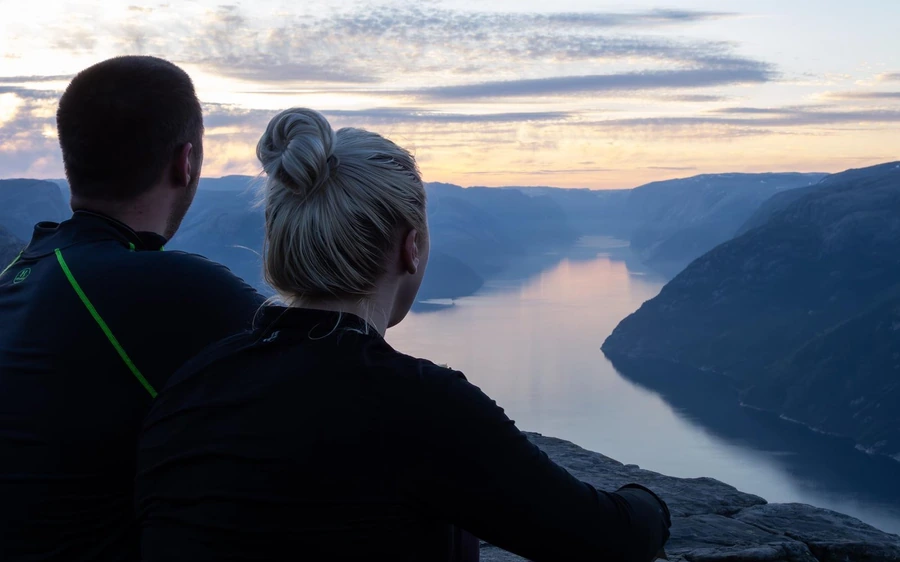 Two hikers seen from behind, looking down a fjord under a pale sky