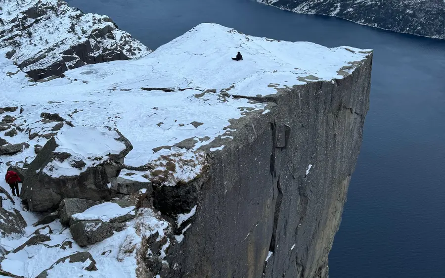 Snow on a flat-topped cliff above dark fjord water, one person sitting on top