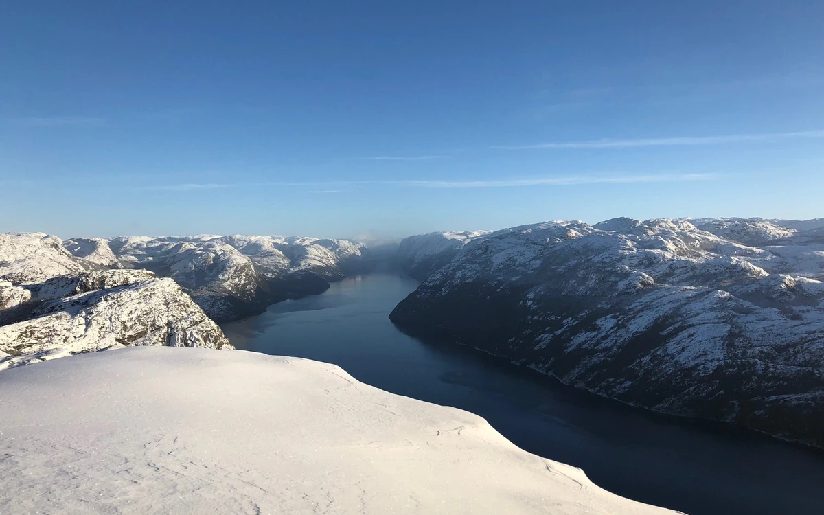 A smooth snow slope in front, the fjord running away between snow-covered ridges under a clear blue sky